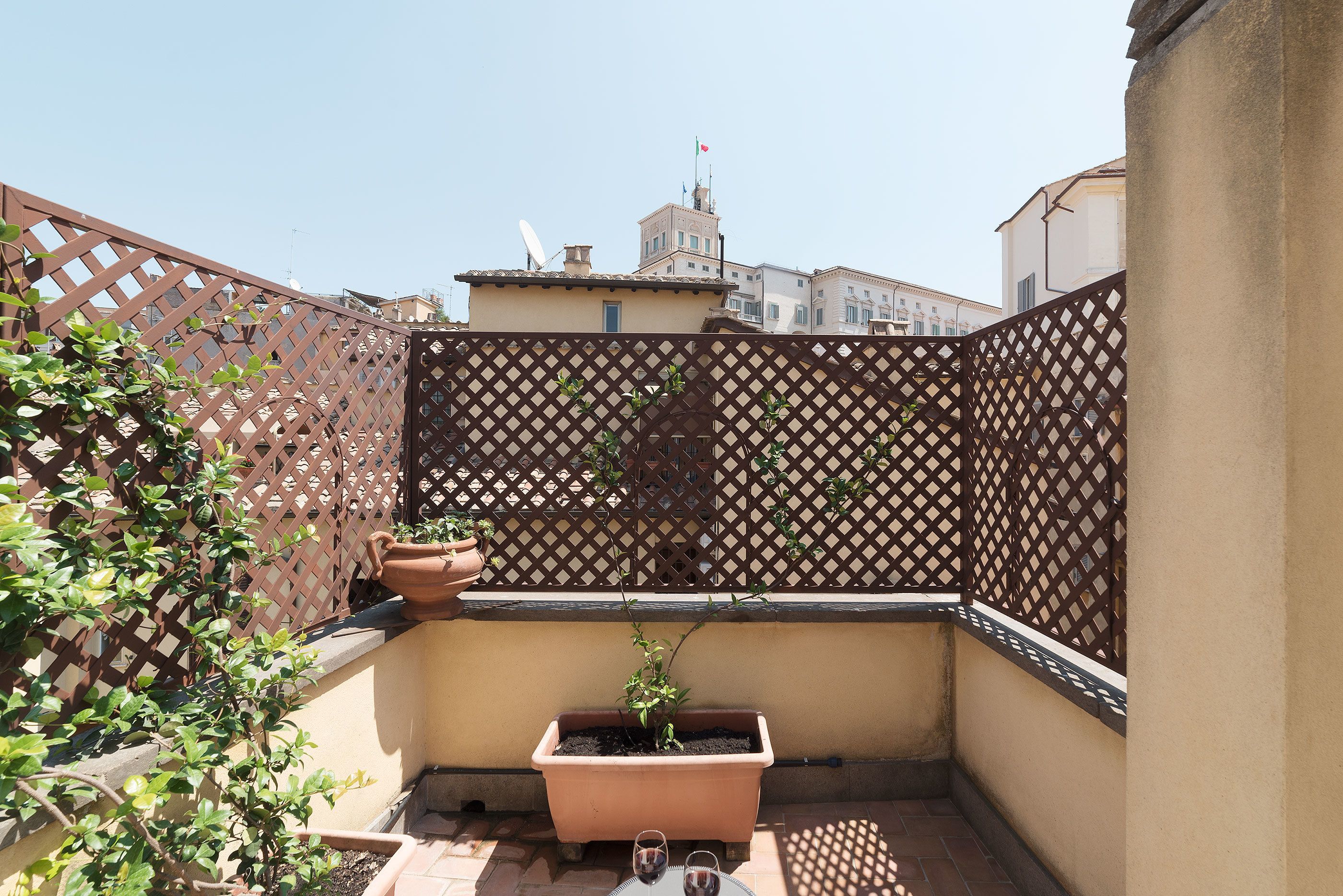 A small outdoor balcony with a lattice fence, a potted plant, and a glass table with two glasses.
