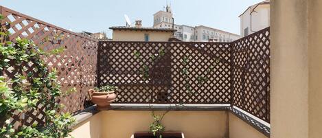 A small outdoor balcony with a lattice fence, a potted plant, and a glass table with two glasses.