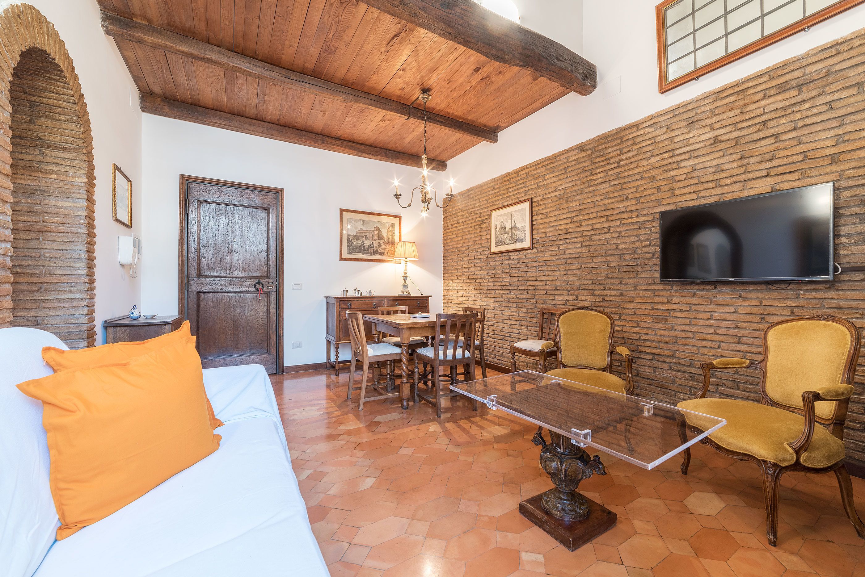 A room with a bed, a dining table with chairs, a TV mounted on a brick wall, and a chandelier.