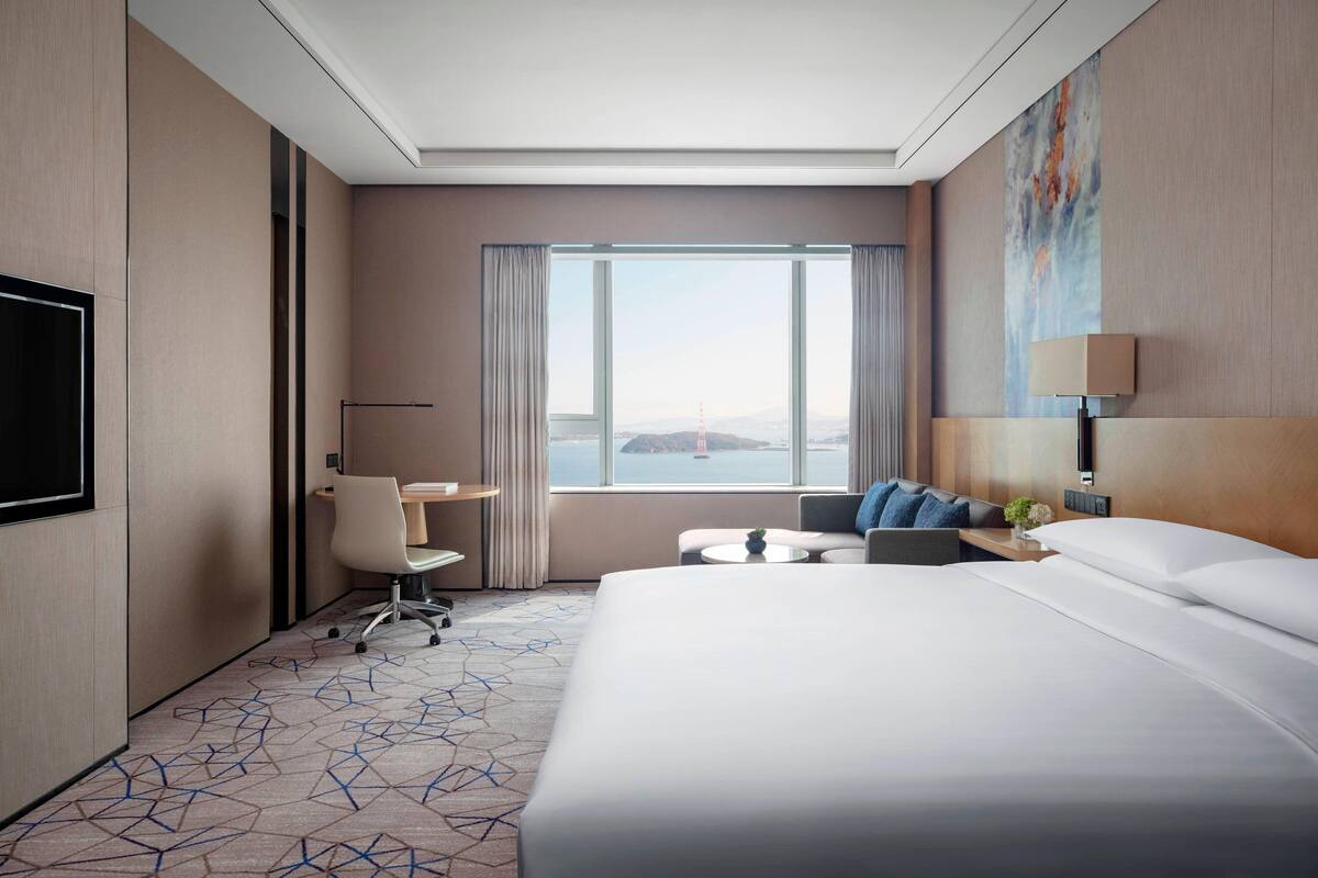 Una habitación de hotel moderna con una cama grande, un escritorio, una silla, un televisor y vistas de la ciudad a través de amplios ventanales.
