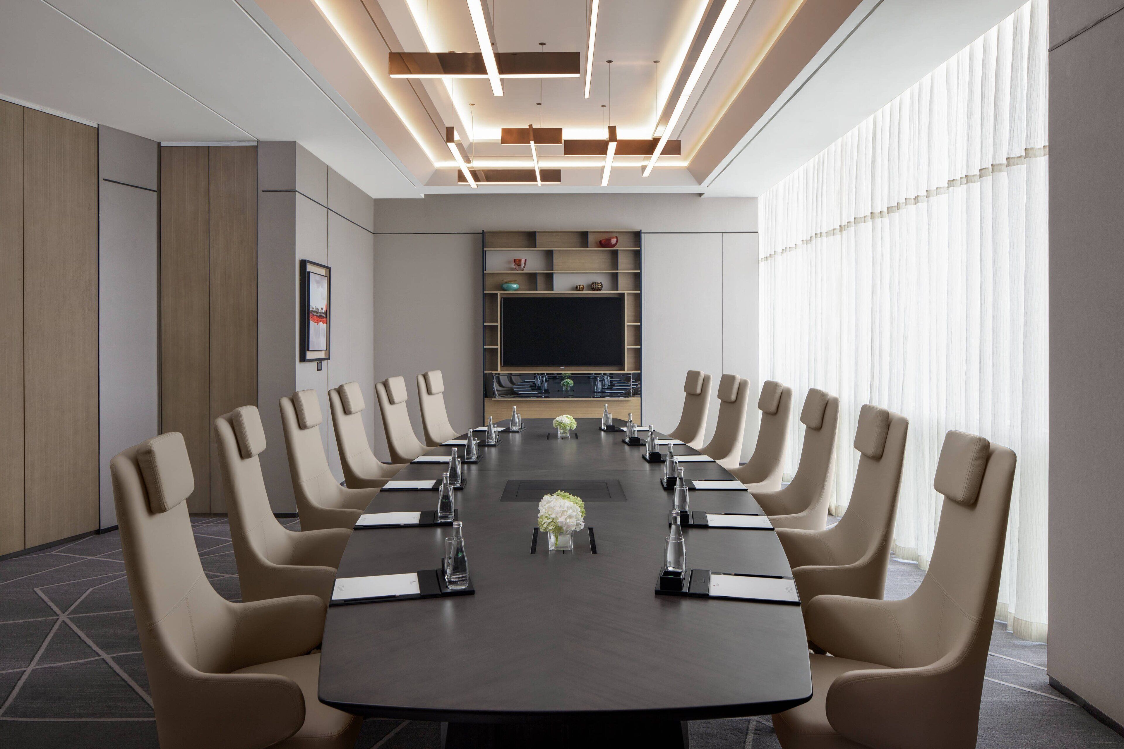 A conference room with a long table, beige chairs, a large screen, and a modern ceiling design.