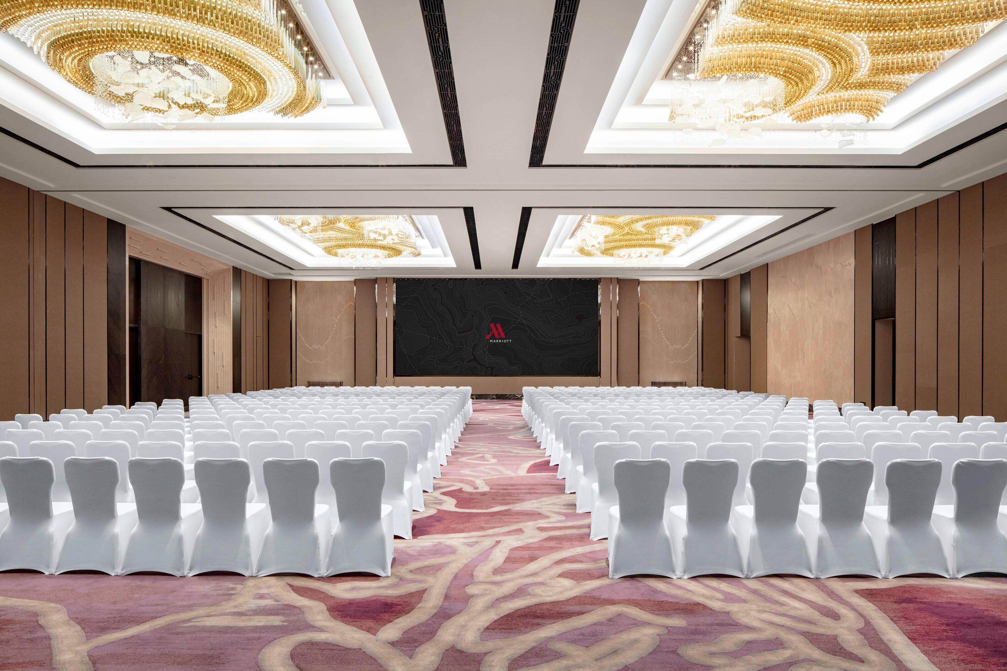 Une salle de conférence avec des rangées de chaises blanches, une moquette rouge et un grand écran à l’avant.