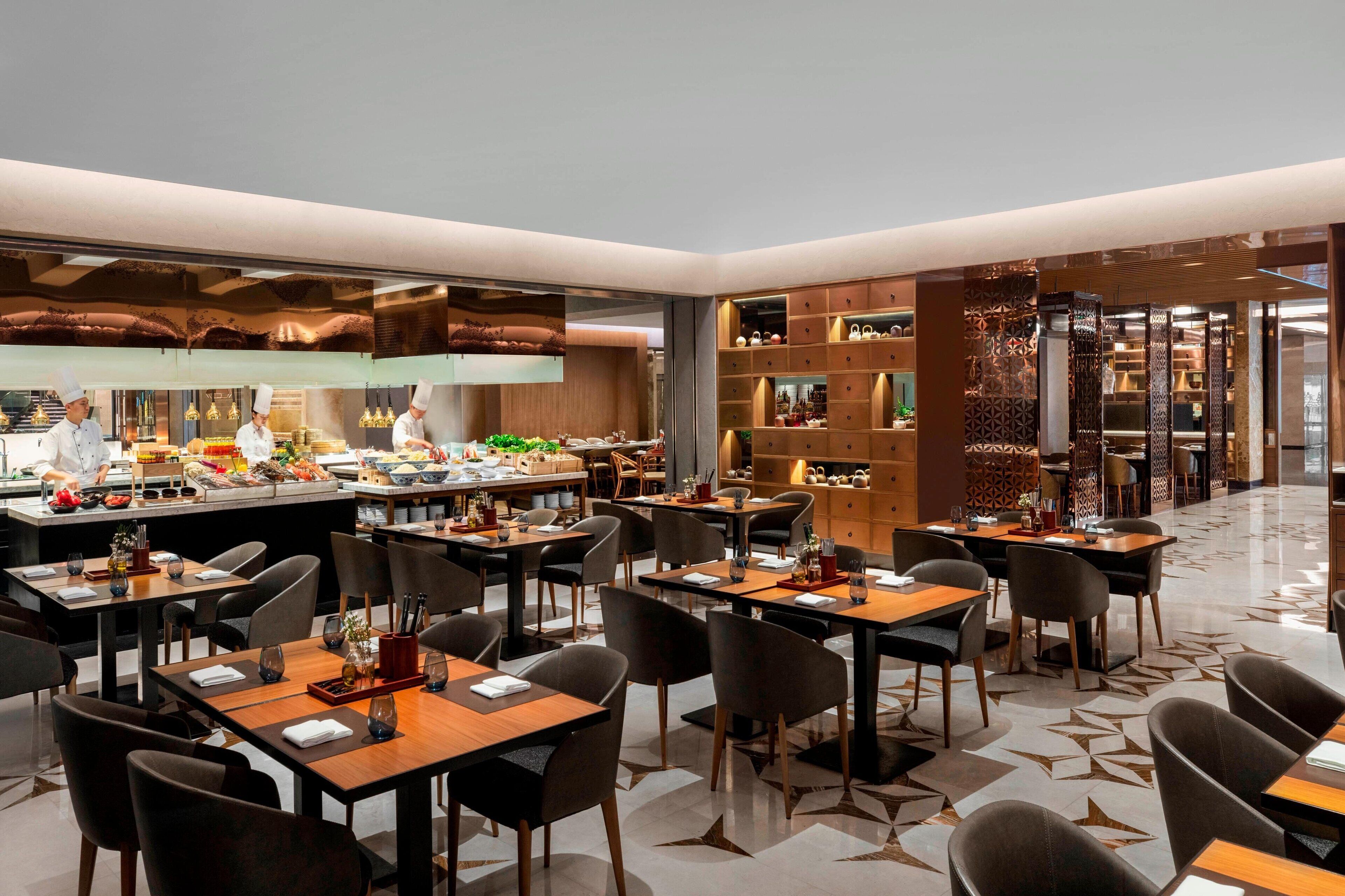 A modern restaurant with multiple dining tables and chairs, a visible kitchen area with chefs, and a patterned floor.