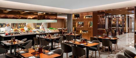 A modern restaurant with multiple dining tables and chairs, a visible kitchen area with chefs, and a patterned floor.