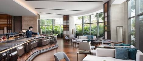 A modern bar area with a curved counter, seating with white and blue cushions, and large windows offering a view of greenery.