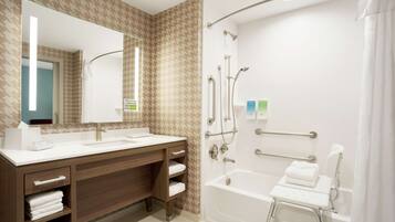 A modern bathroom with a wooden vanity, a large mirror, a bathtub with a showerhead, and a handrail.