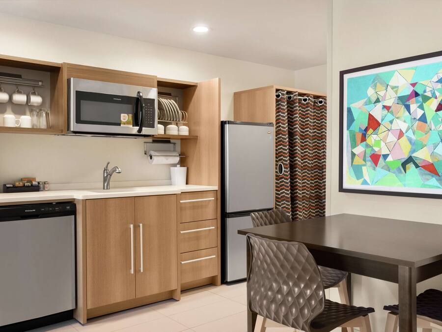 A compact kitchen with a microwave, refrigerator, and dishwasher. There is a dining table with chairs and a framed abstract painting on the wall.