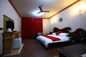 A hotel room with a bed, a desk, a chair, a television, a ceiling fan, and a red curtain. - Hotel Anandam (Gurugram)
