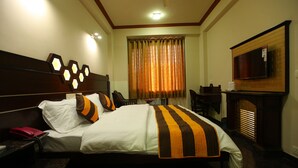 A hotel room with a large bed, a TV mounted on the wall, a desk with a chair, and a fireplace. - Hotel Anandam (Gurugram)