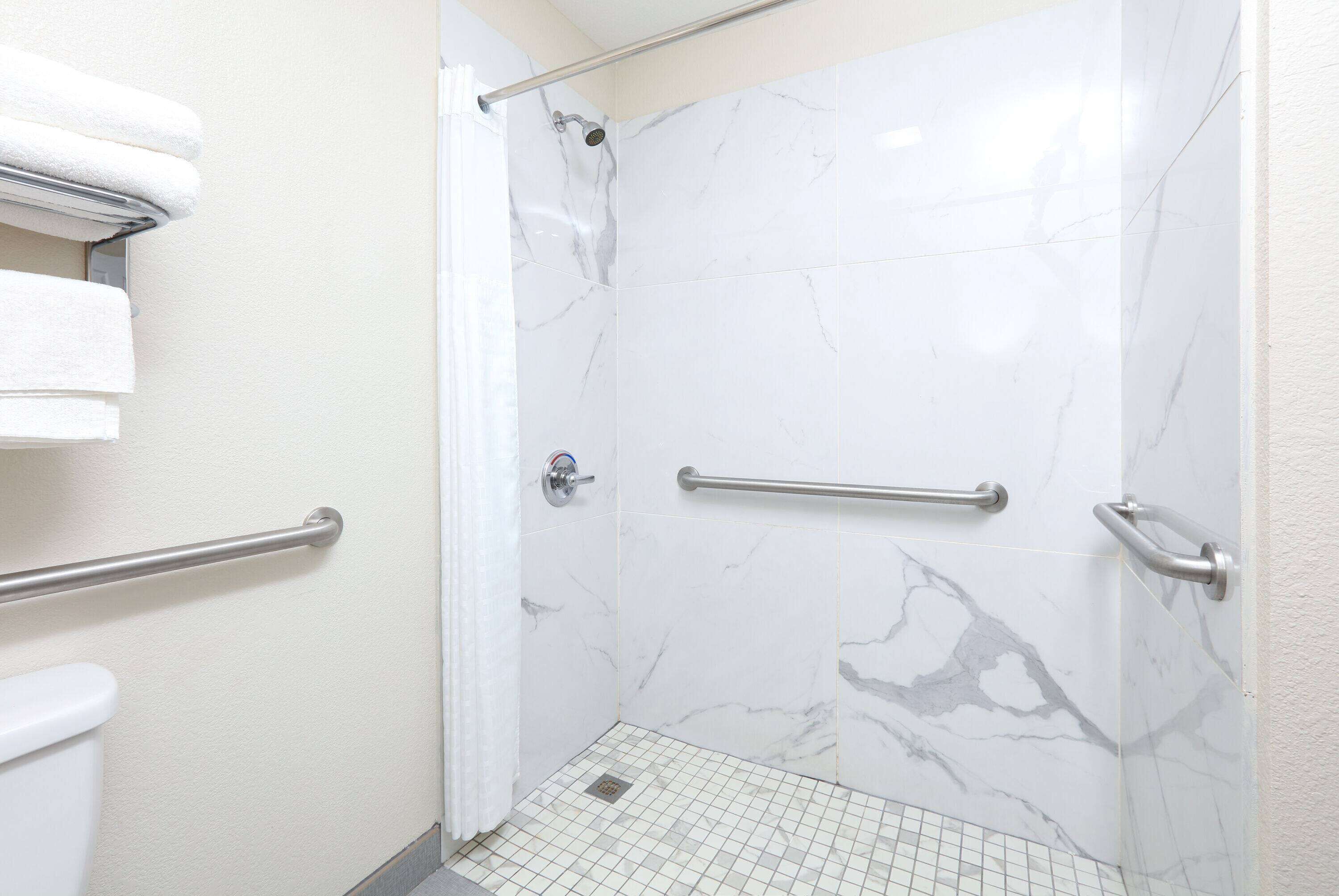 Une salle de bain aux murs recouverts de marbre blanc, une douche avec un pommeau de douche amovible et une barre d’appui pour plus de sécurité.