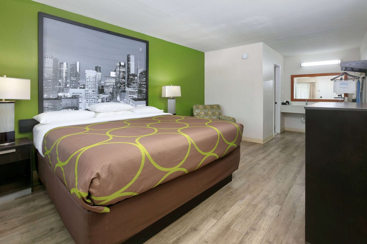 Ett hotellrum med en säng, gröna väggar, en målning som föreställer en stadsbild, ett skrivbord med en lampa och en spegel.
