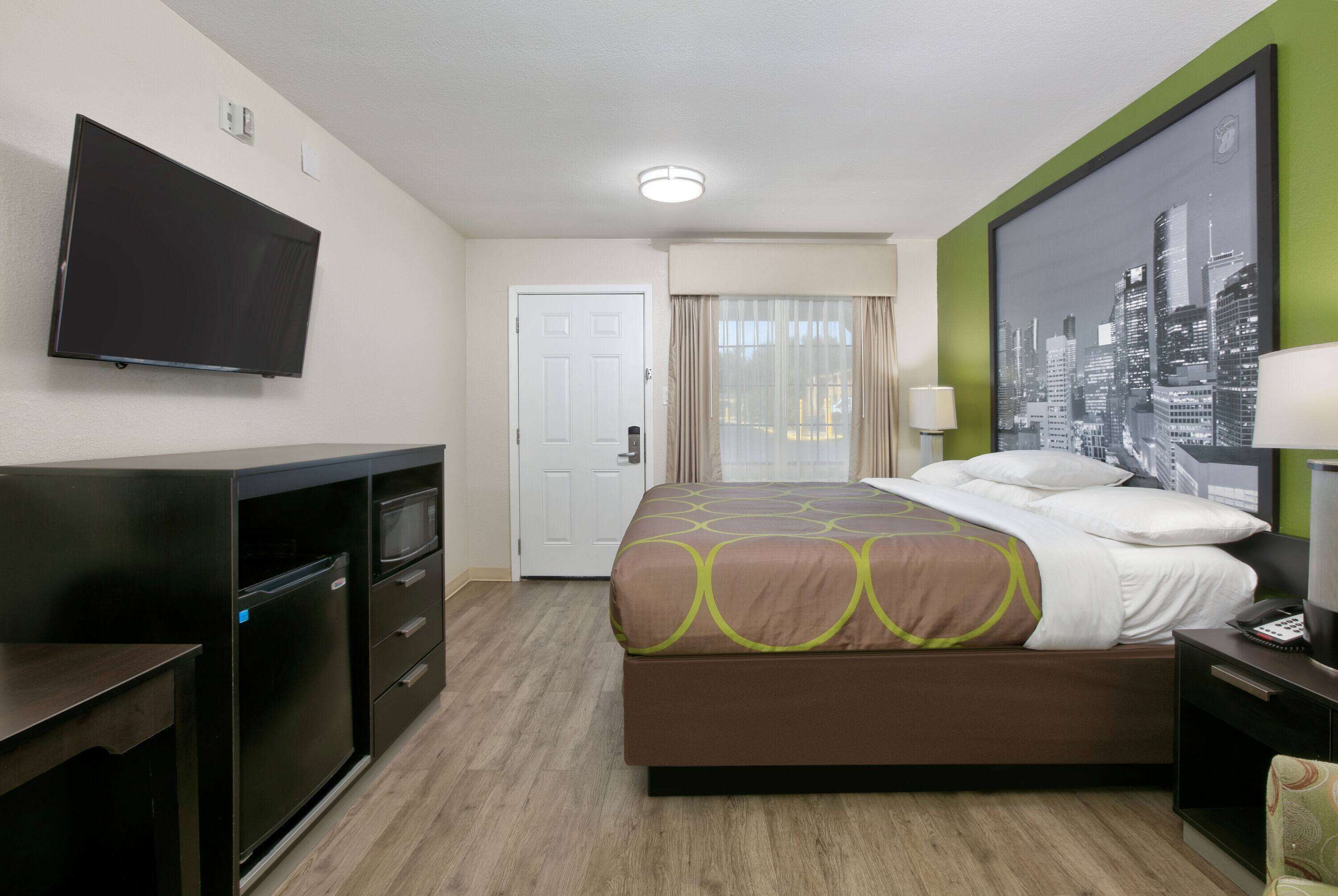 A hotel room with a bed, a desk, a flat-screen TV, a door, a window with curtains, and a cityscape painting.