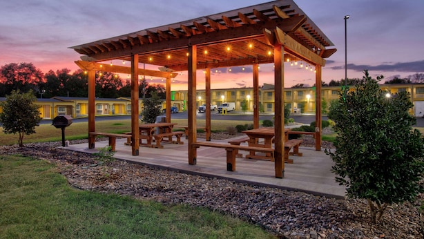 A covered outdoor seating area with picnic tables and string lights, surrounded by a landscaped garden with a building in the background. - Super 8 by Wyndham Palestine (Palestine)