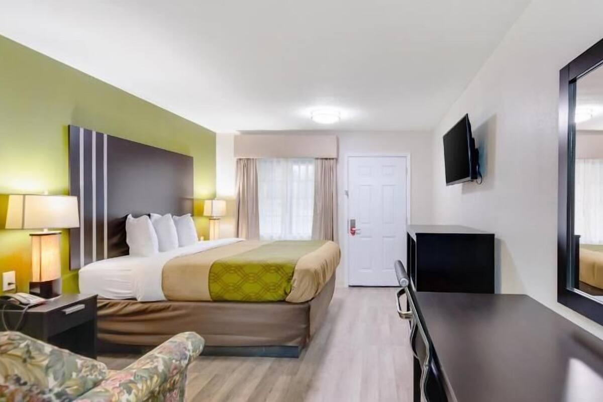 Ett hotellrum med en säng, ett skrivbord, en stol, en tv och en dörr.