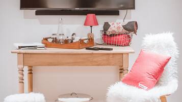 Ein modernes Wohnzimmer mit Flachbildfernseher an der Wand, einem Holztisch mit Lampe, einem Sessel mit flauschigem weißen Kissen und einem kleinen weißen Hocker.