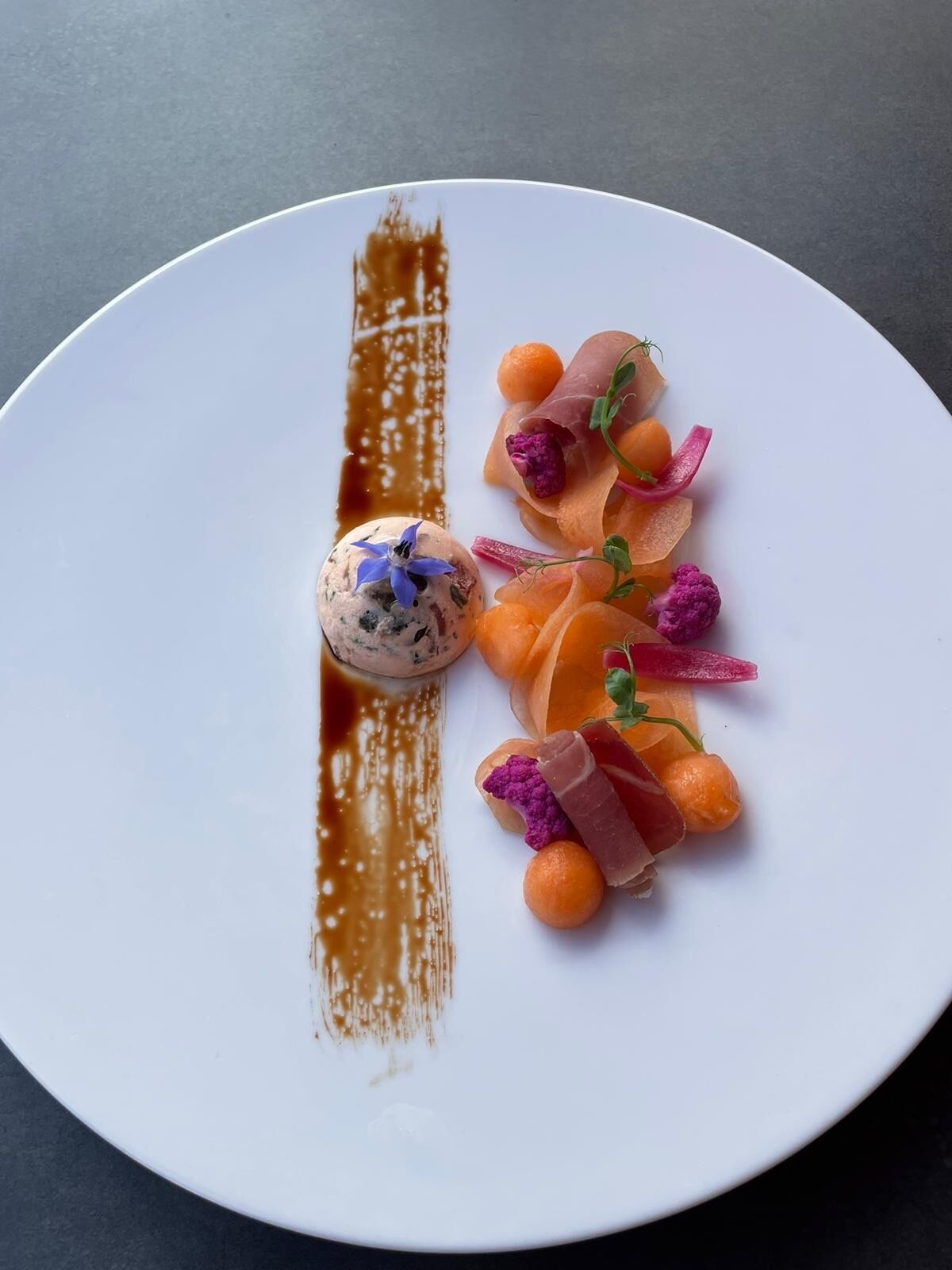 Un plato blanco con un postre que incluye una porción esférica, esferas anaranjadas y elementos rosados y morados, sobre una superficie oscura.