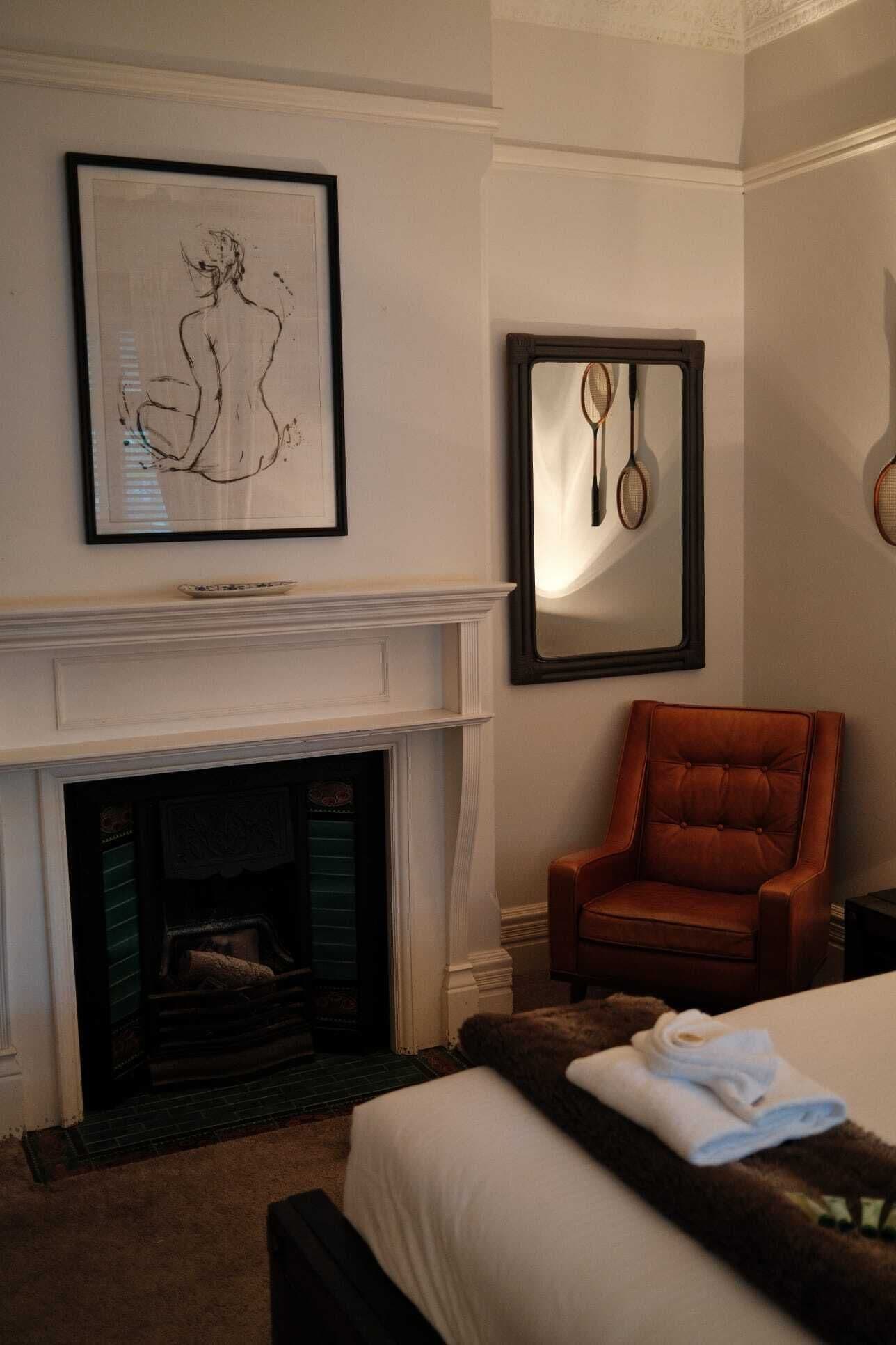 A hotel room with a fireplace, a framed artwork, a mirror with tennis rackets, a leather armchair, and a neatly made bed.
