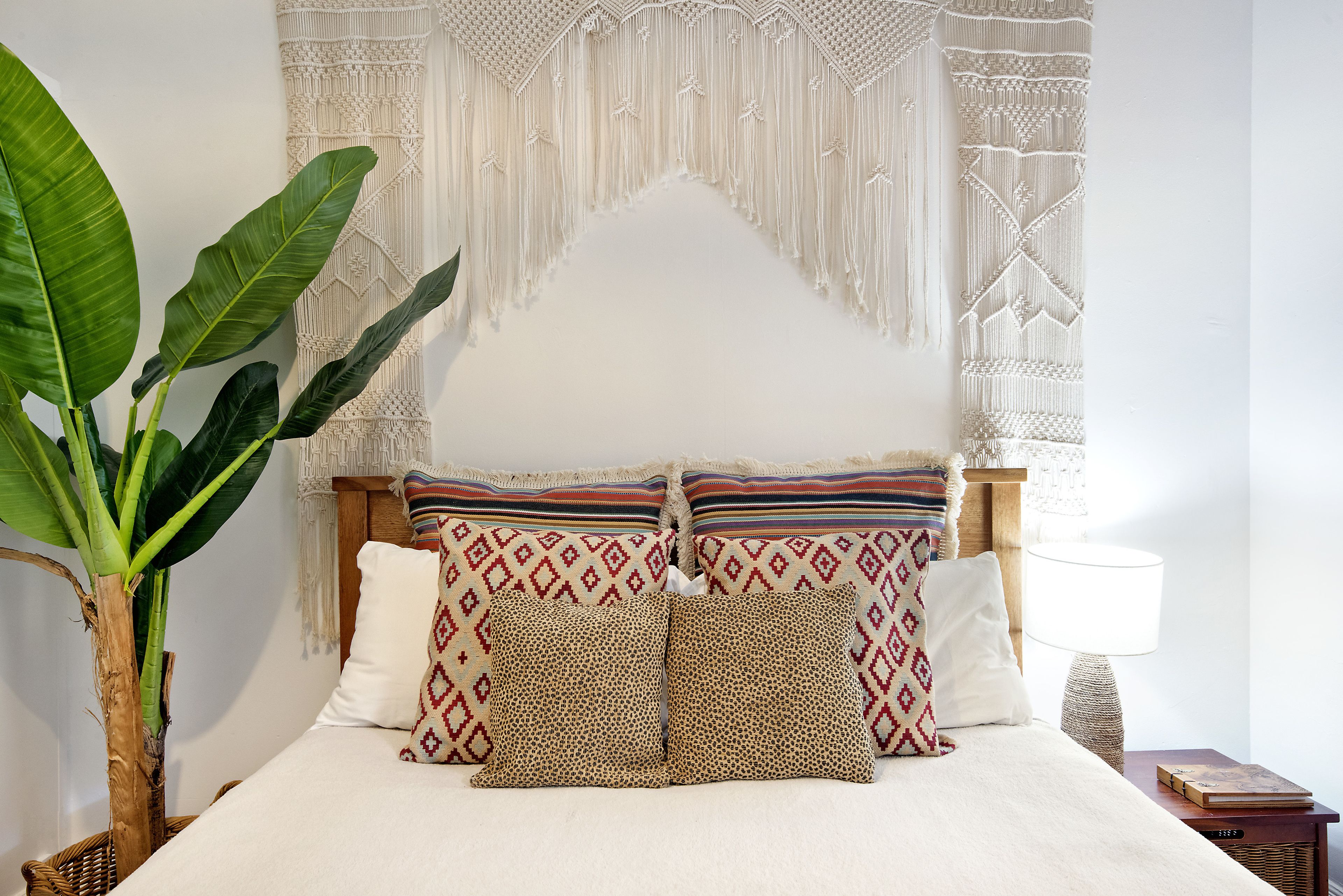 A neatly made bed with patterned pillows, a macrame wall hanging, and a large plant beside the bed.