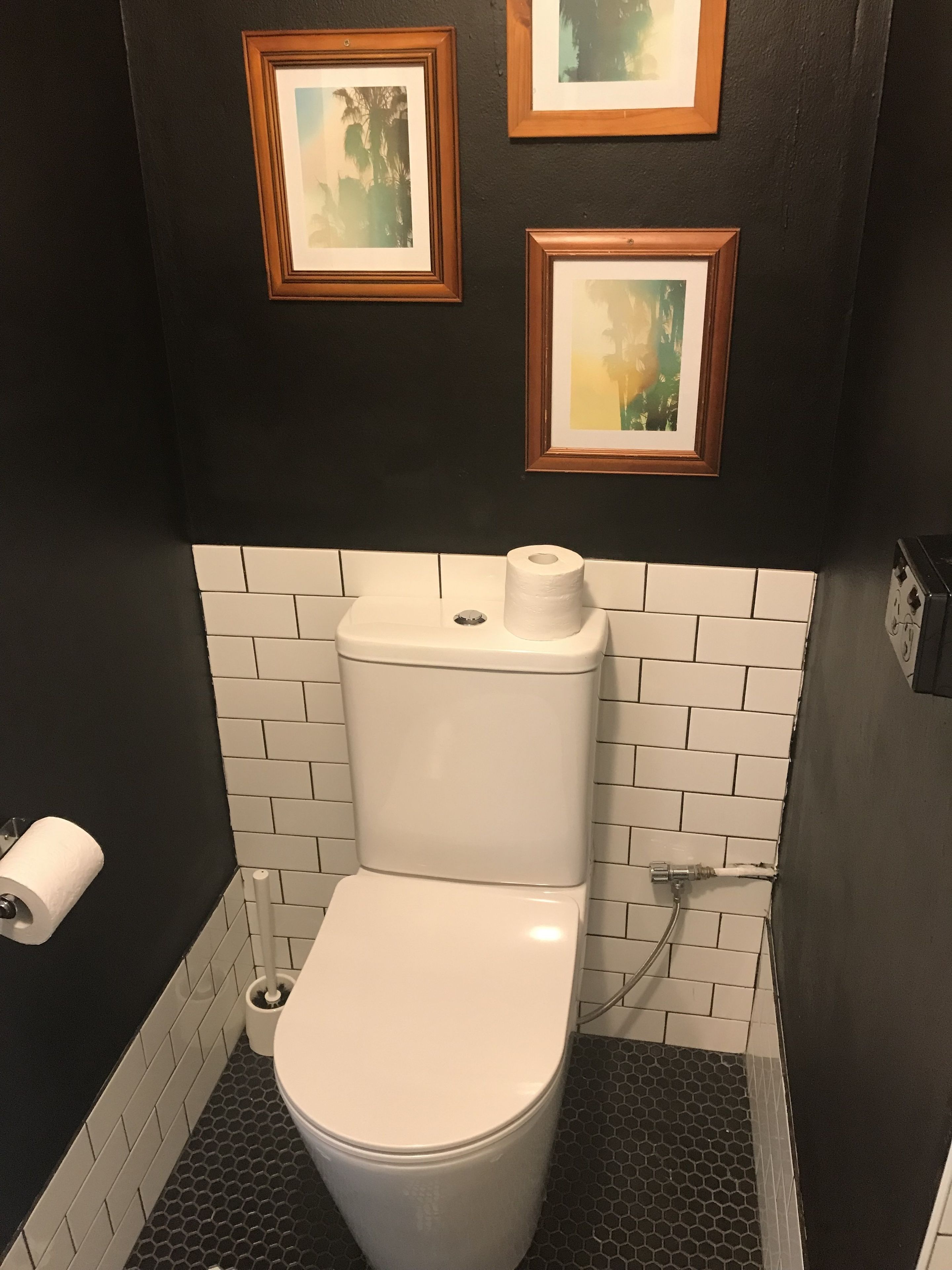 A small bathroom with a white toilet, black and white tiled walls, and two framed pictures on the wall.