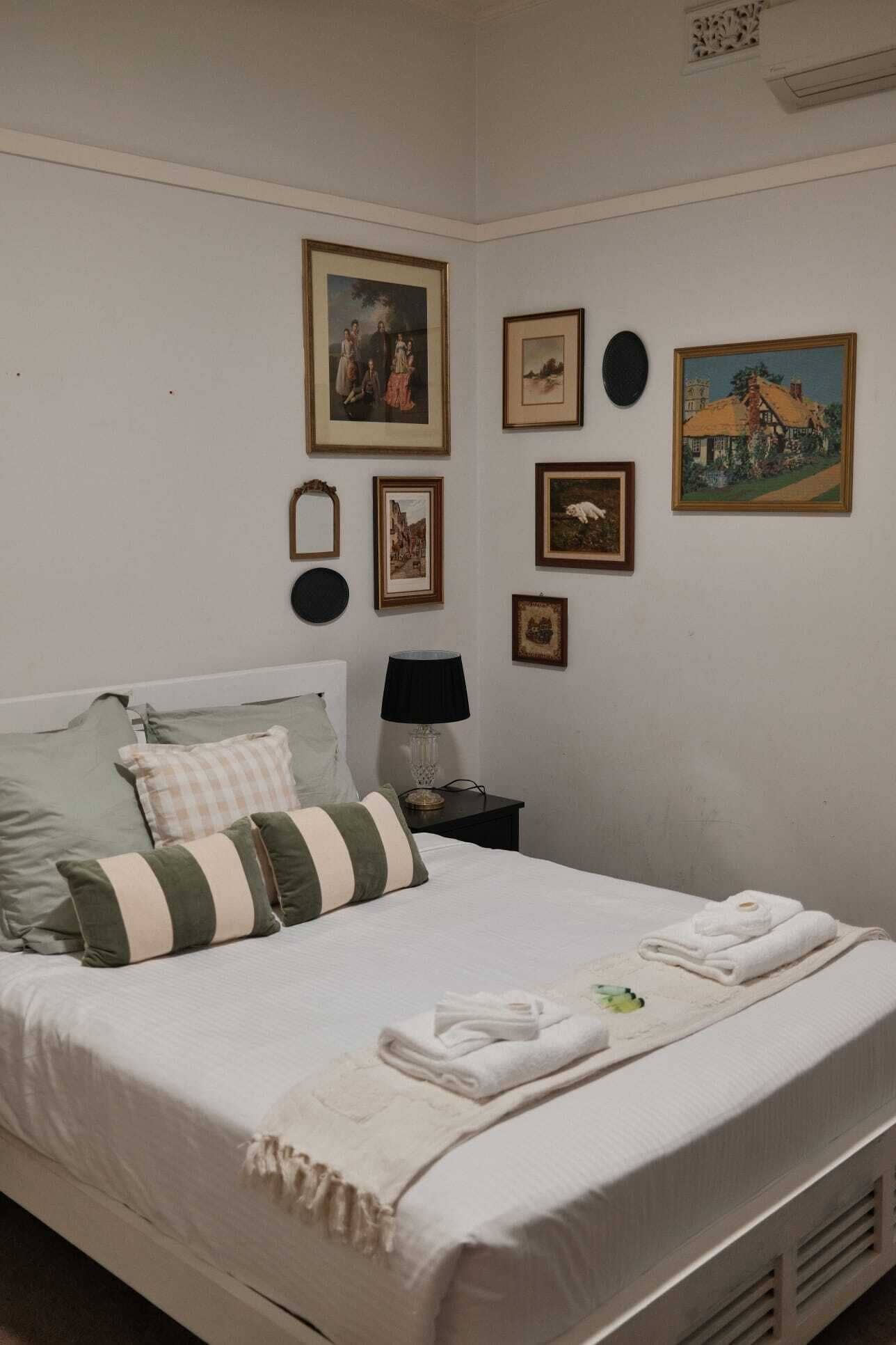 Un dormitorio con cama, almohadas, una mesita de noche con lámpara y cuadros en la pared.
