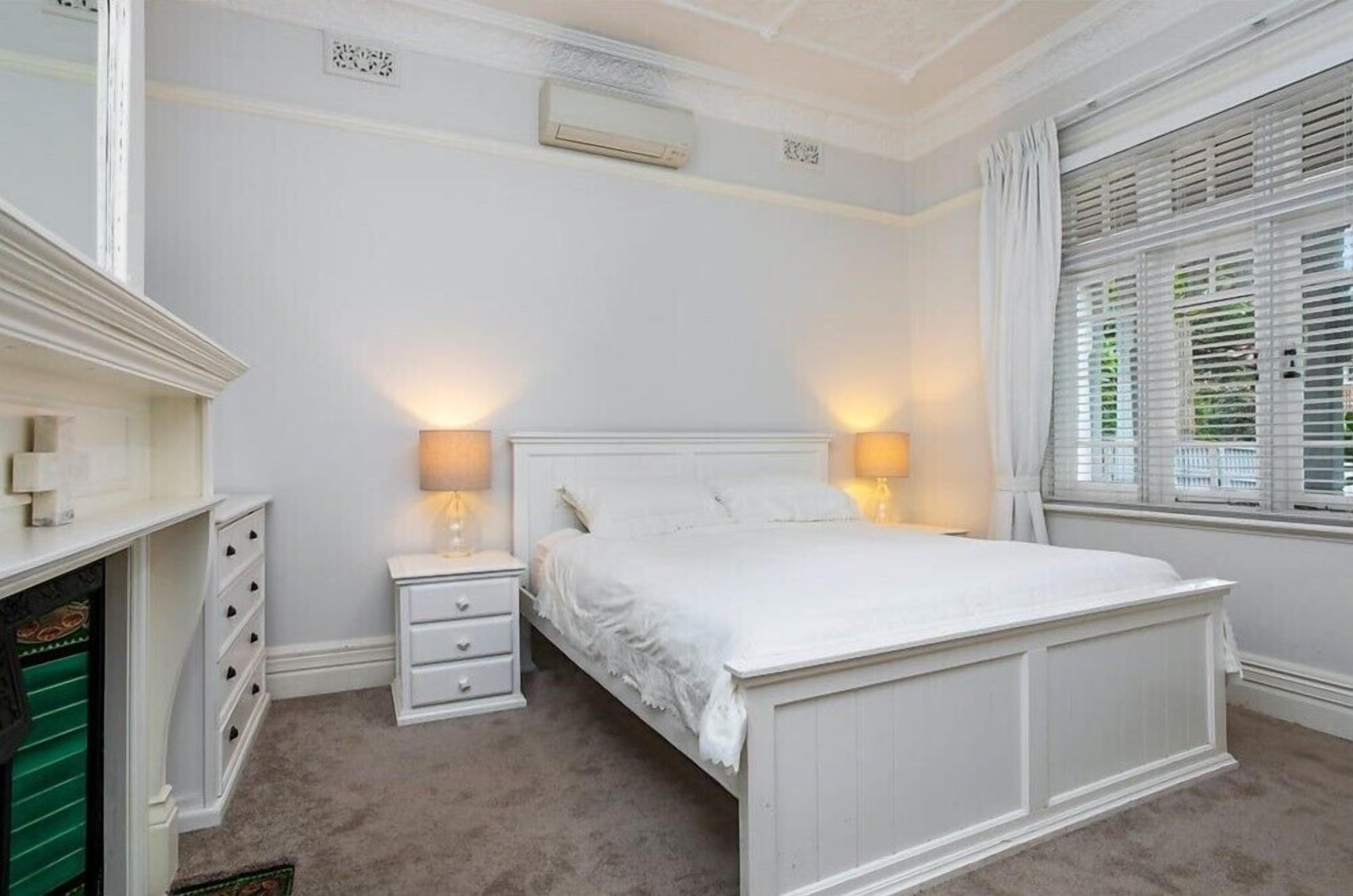 A bedroom with a white bed, two bedside tables with lamps, a dresser, and a window with blinds.