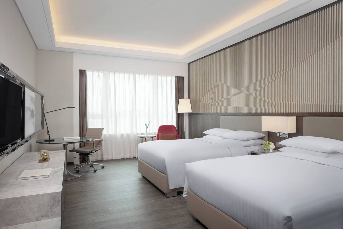 Une chambre d’hôtel avec deux lits, un bureau doté d’un téléviseur à écran plat, une chaise, une lampe et une vue sur l’extérieur grâce à de grandes fenêtres.