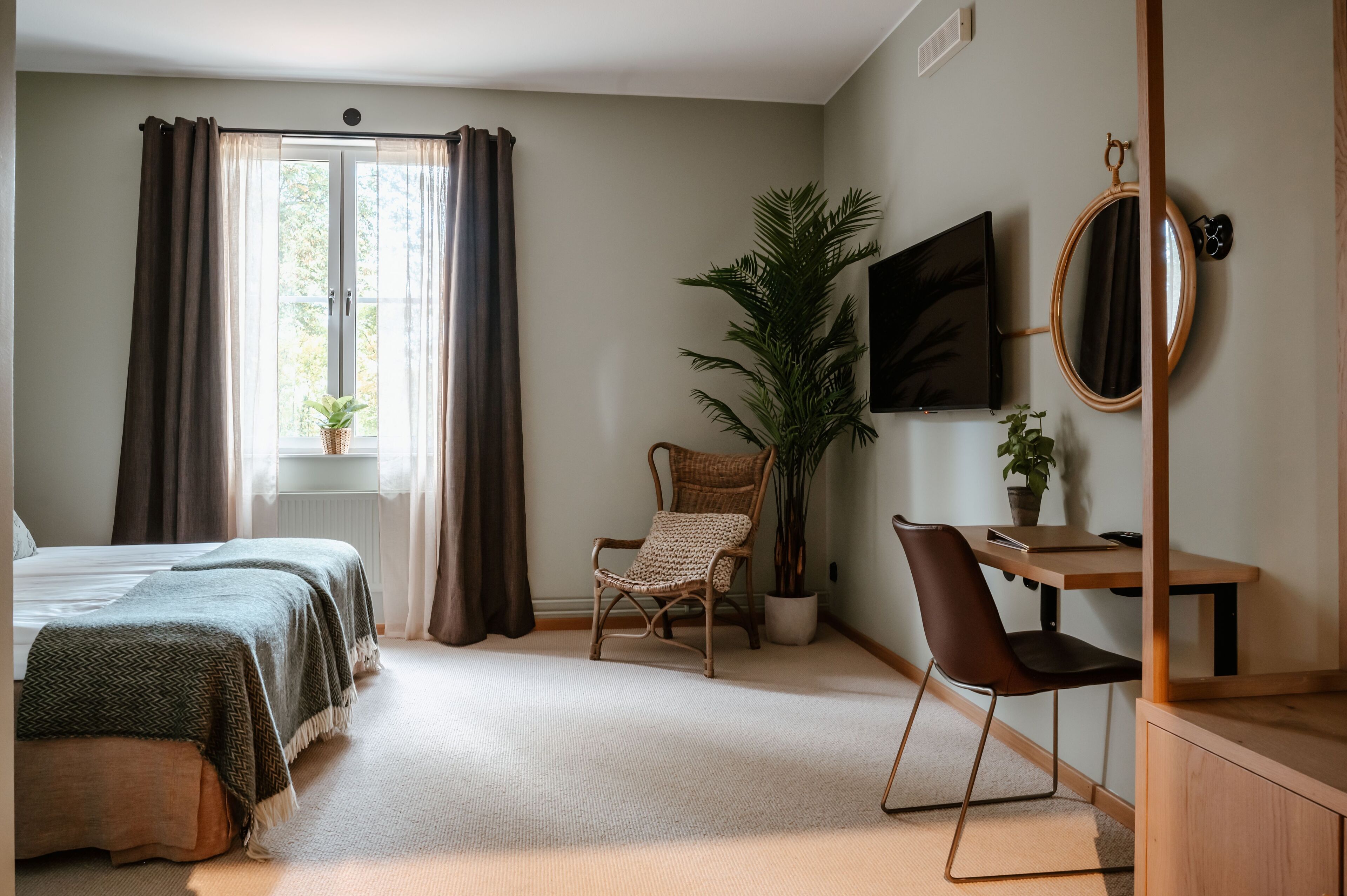 Ein Schlafzimmer mit einem Bett, zwei Nachttischen, einem Schreibtisch mit Stuhl, einer großen Kübelpflanze, einem an der Wand montierten Fernseher und einem Spiegel.