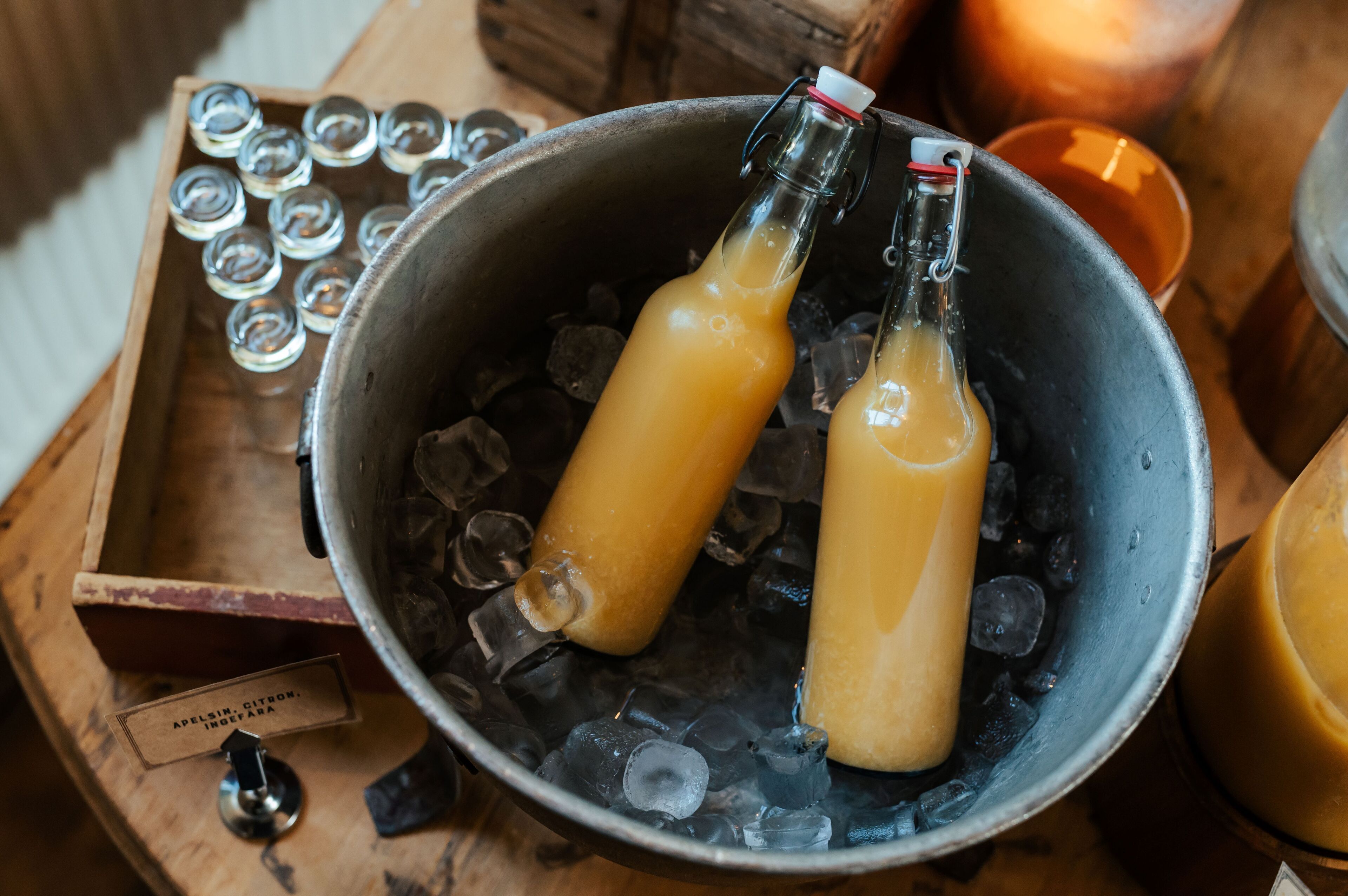 Two bottles of amber liquid in a metal bucket filled with ice cubes, with a wooden crate and a metal container in the background.