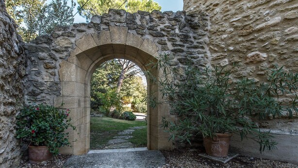 Un portail en pierre en forme d’arc donne accès à un jardin agrémenté de plantes en pot et d’un chemin de gravier.