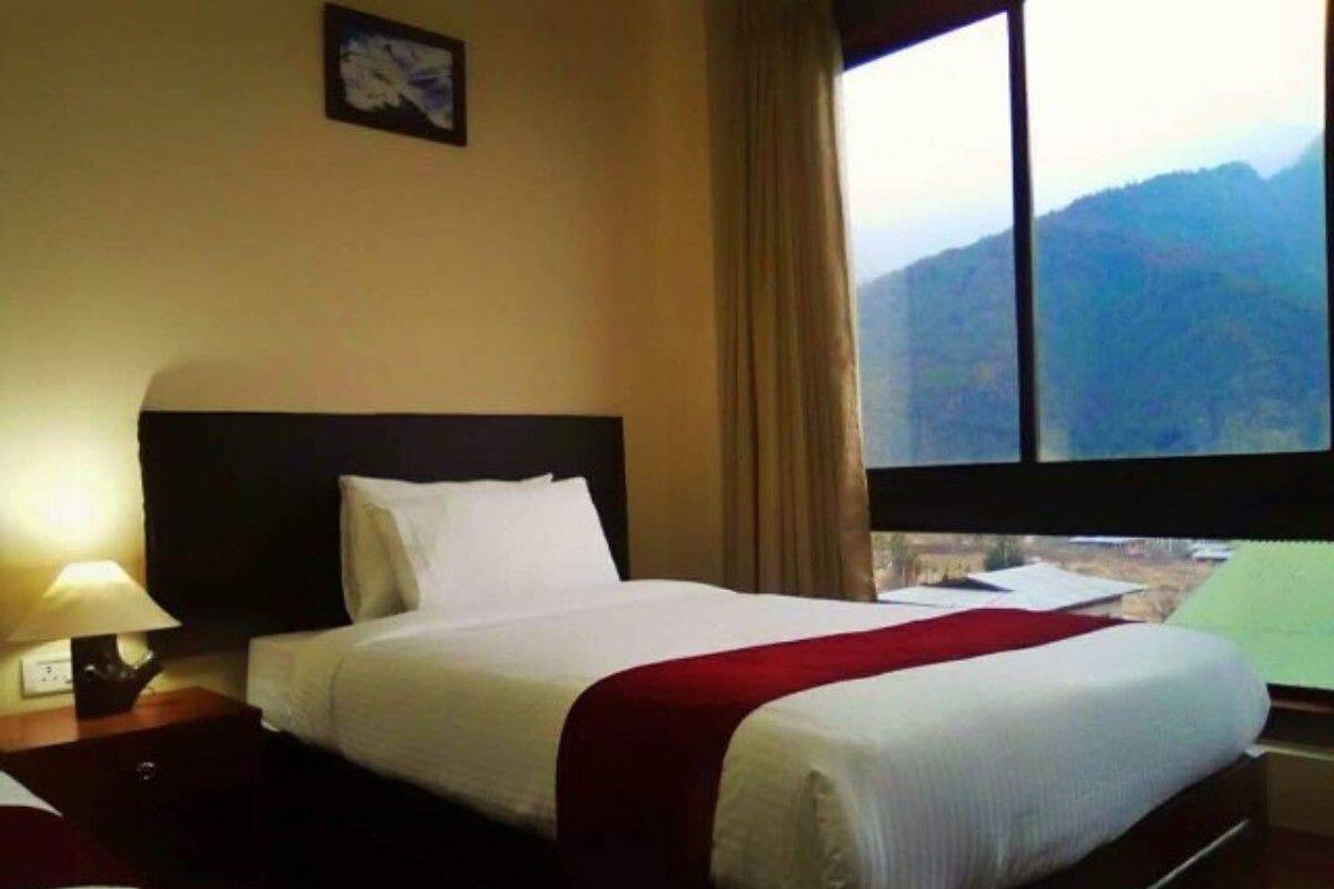 Habitación de hotel con una cama, una mesita de noche con lámpara, una ventana con vista a las montañas y un cuadro en la pared.