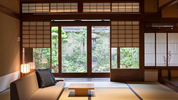 A traditional Japanese room with tatami flooring, a low wooden table, a sofa with cushions, and sliding doors leading to a garden. - HANARé Genmachi (Kyoto)