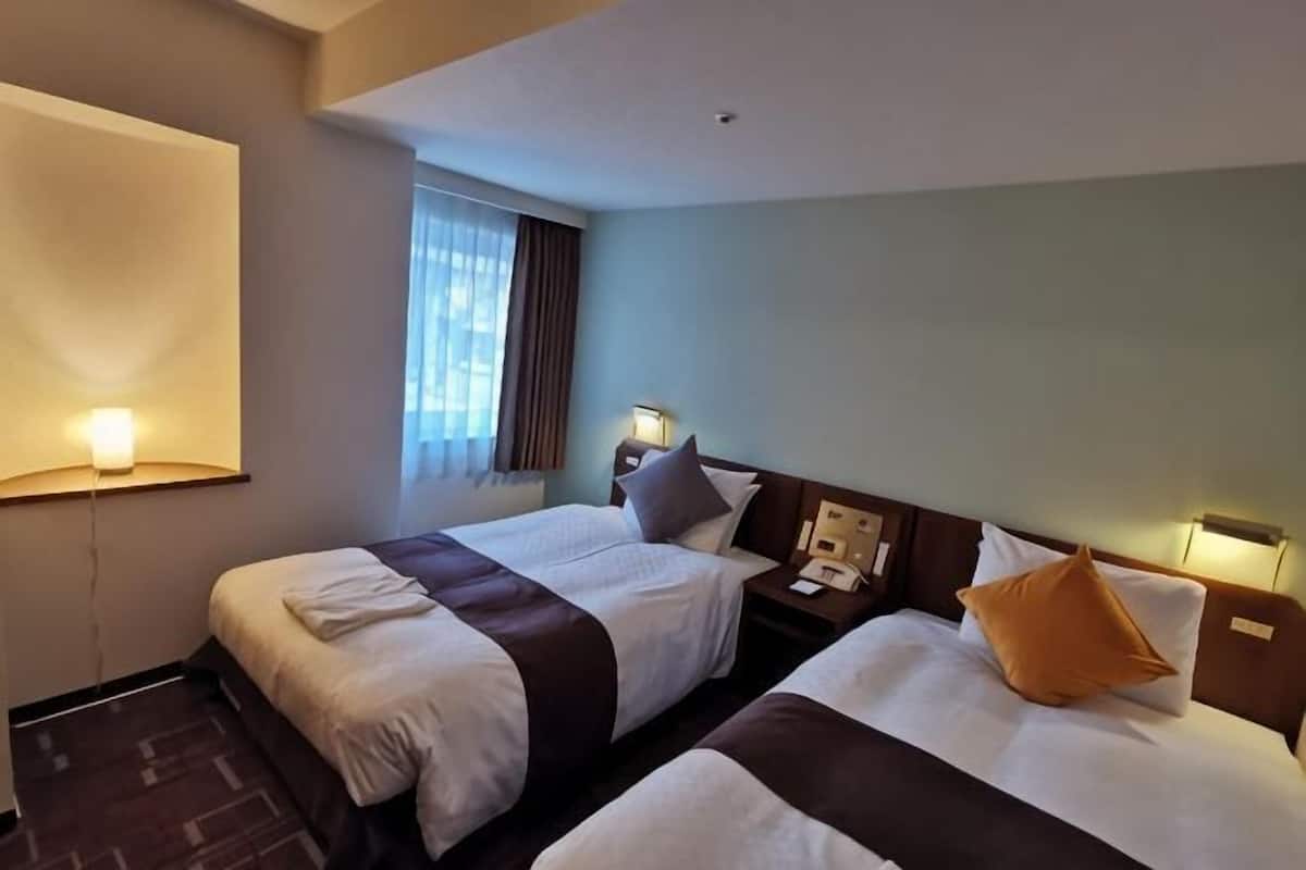 A hotel room with two beds, a window with curtains, a nightstand with a lamp, and a wall-mounted phone.