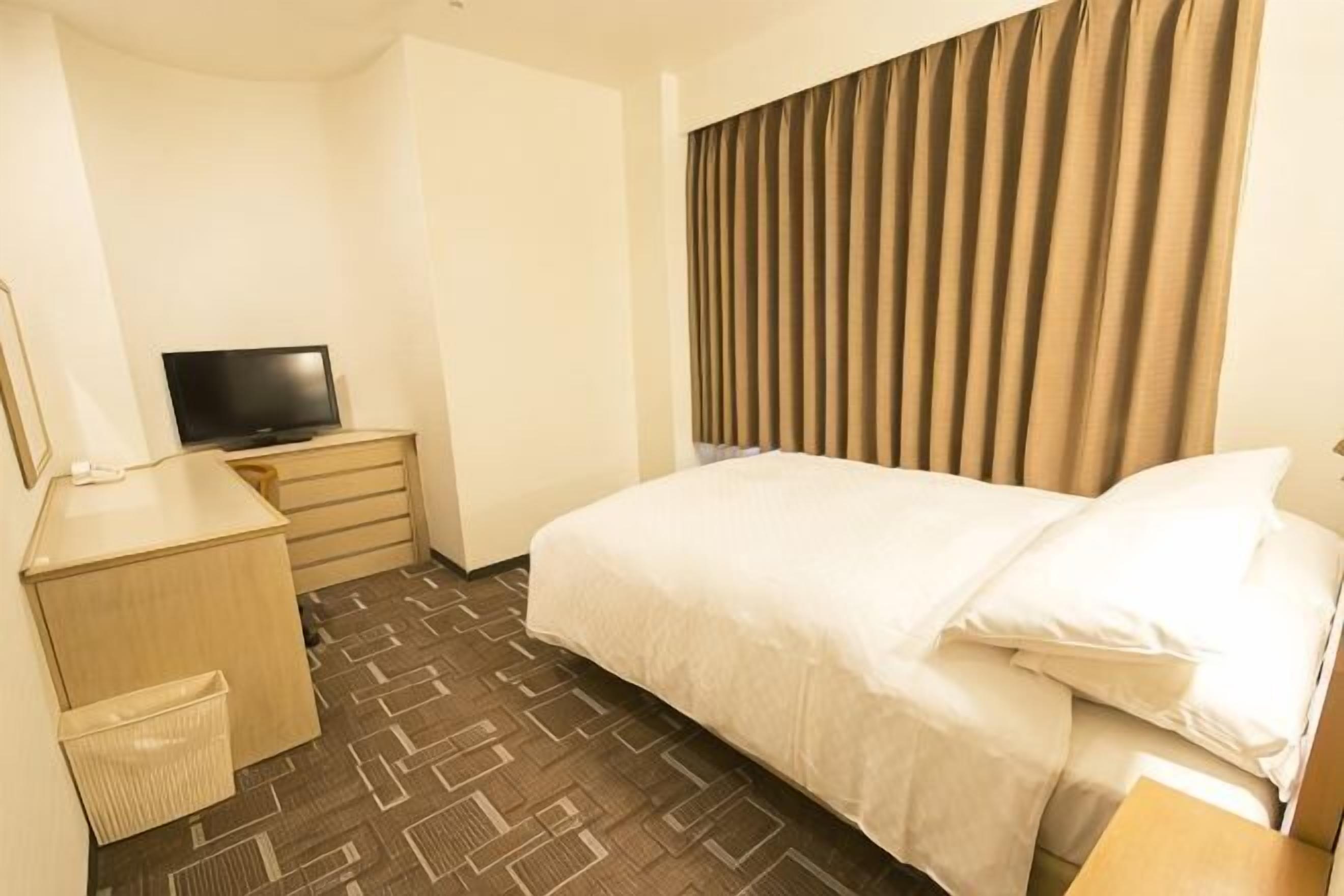 Une chambre d’hôtel comprenant un lit, un bureau avec une télévision, une petite commode et une corbeille à déchets.