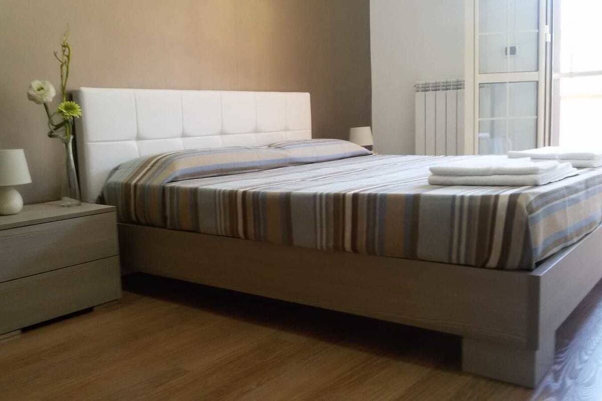 A modern bedroom with a large bed, a nightstand with a lamp, and a vase with flowers.