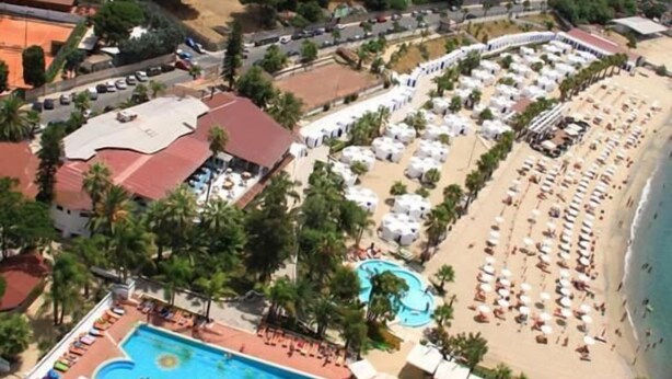 Luchtbeeld van een resort met een zwembad, een strandgedeelte met parasols en een weg.