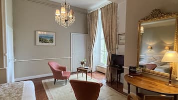 A hotel room with a bed, a sitting area with a chair, a small table with a vase, a desk with a computer, and a large mirror.