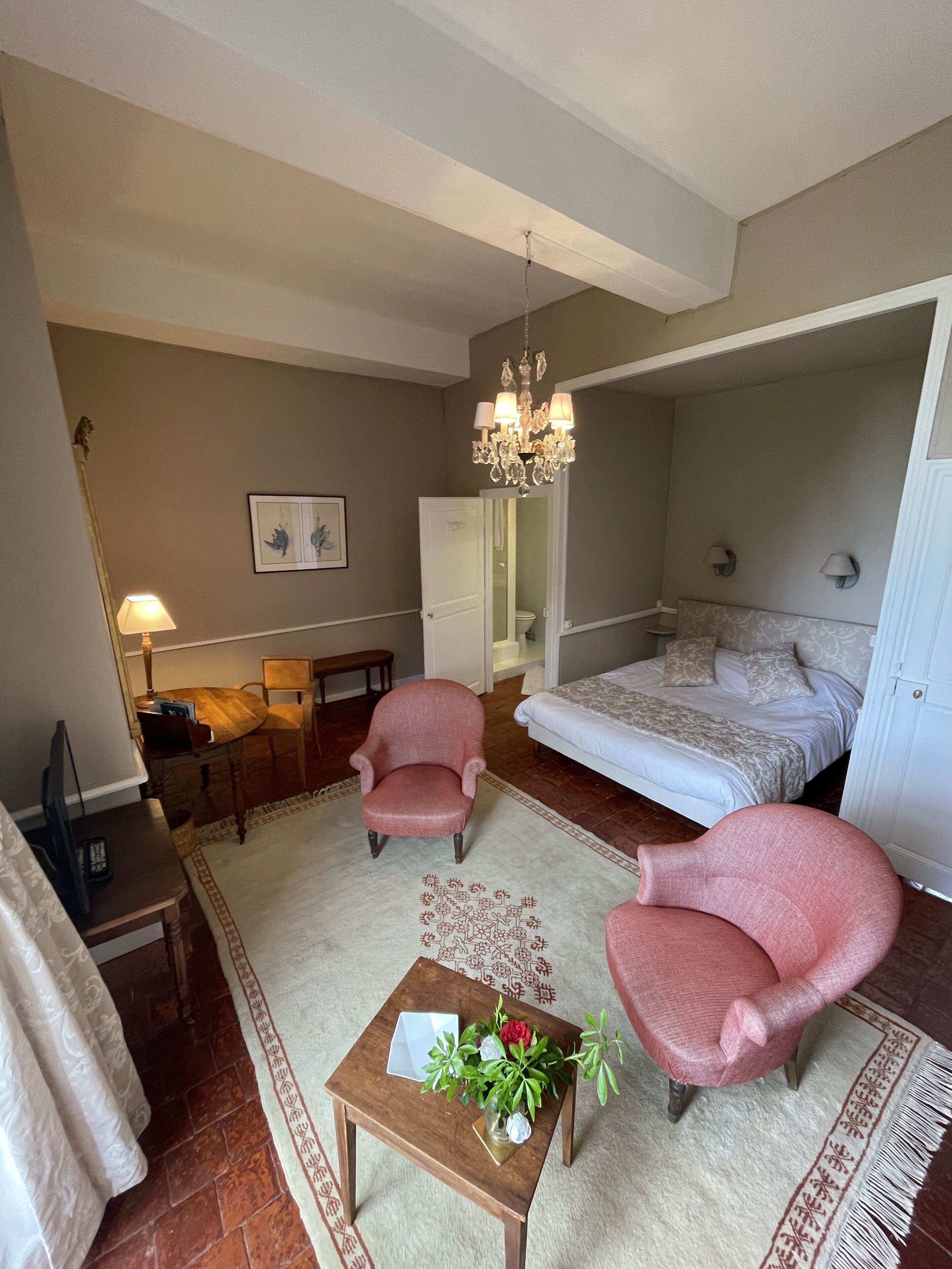 Une chambre à coucher avec un lit, deux fauteuils, une petite table sur laquelle est posé un vase de fleurs, une lampe et un tableau accroché au mur.