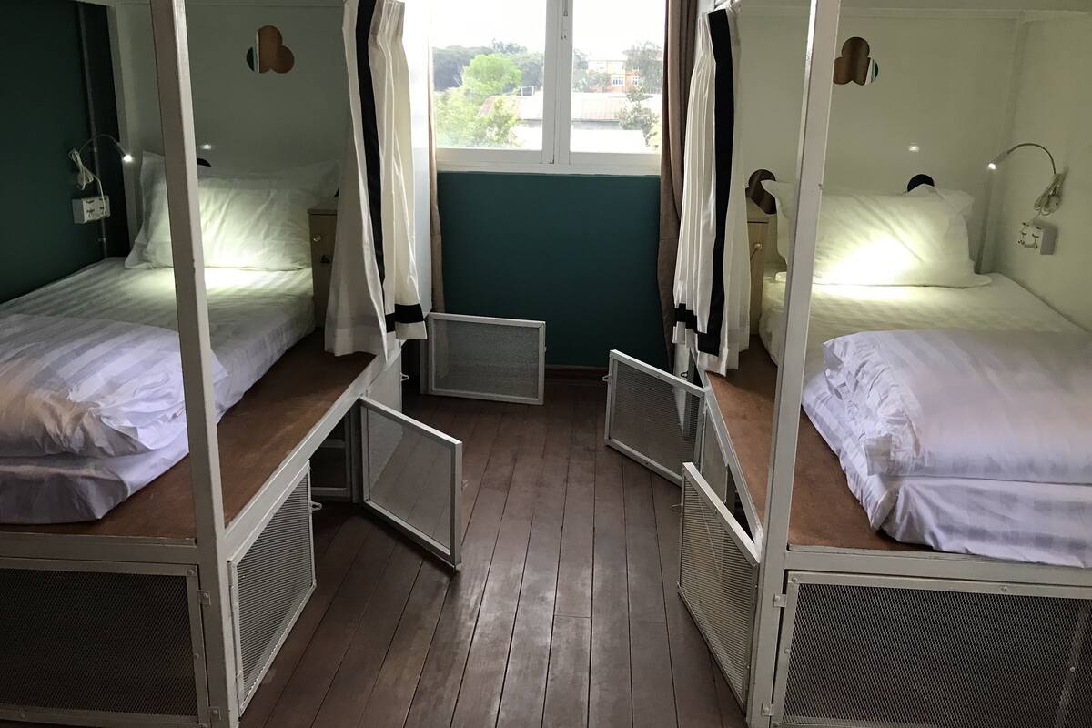 A room with bunk beds, a window with curtains, a bedside table, and a wall-mounted light fixture.