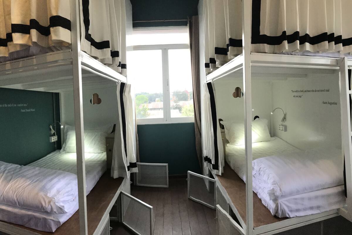 A room with two bunk beds, each with a small bedside table and a lamp. There is a window with curtains, and a quote by Saint Augustine is visible on the wall.