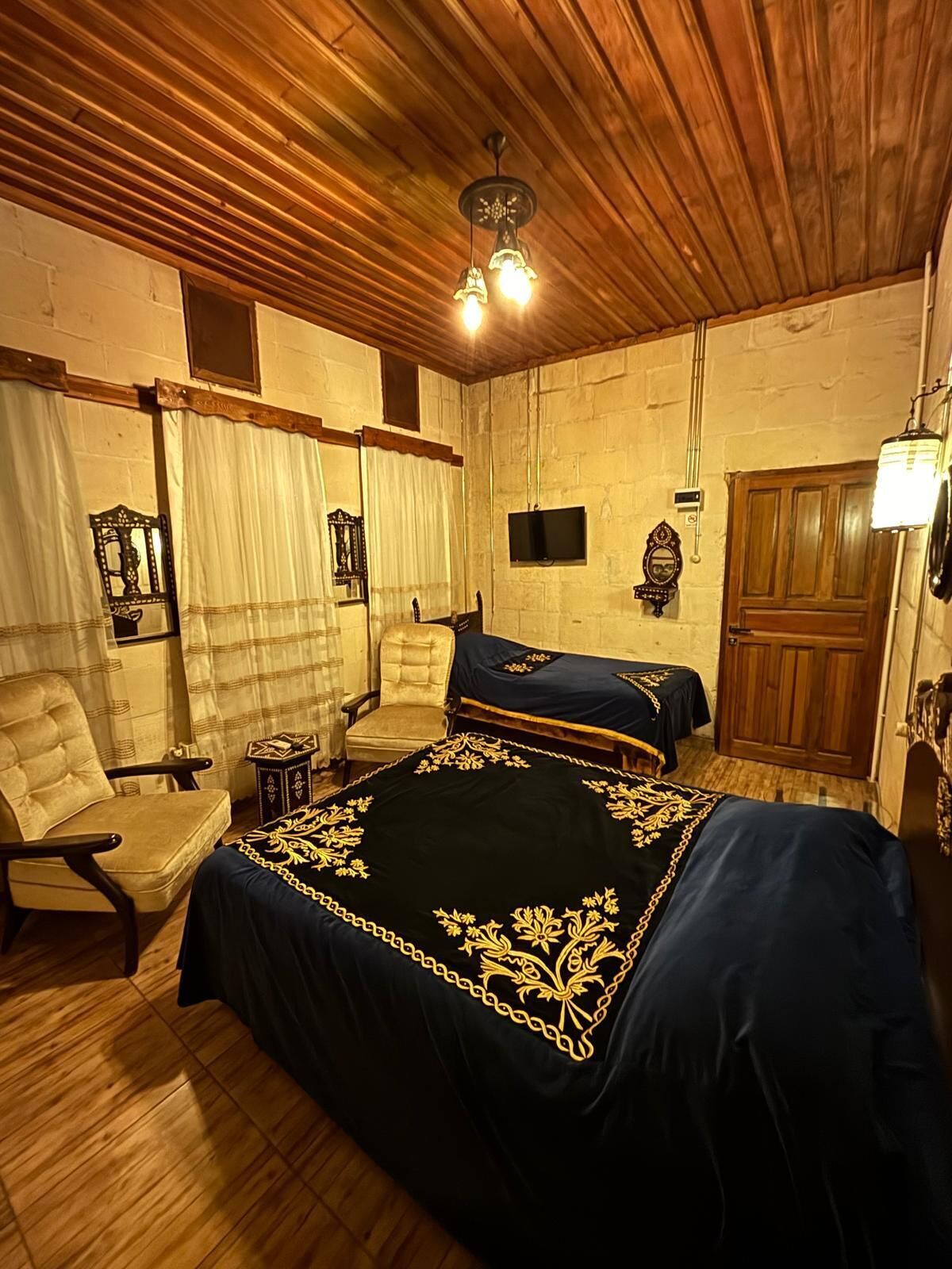 A room with a large bed, a chair, a wooden ceiling, and a television on the wall.