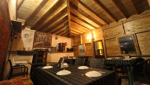 Un coin repas rustique, avec des tables en bois disposées pour accueillir quatre personnes, des poutres en bois au plafond et un espace cuisine en arrière-plan.