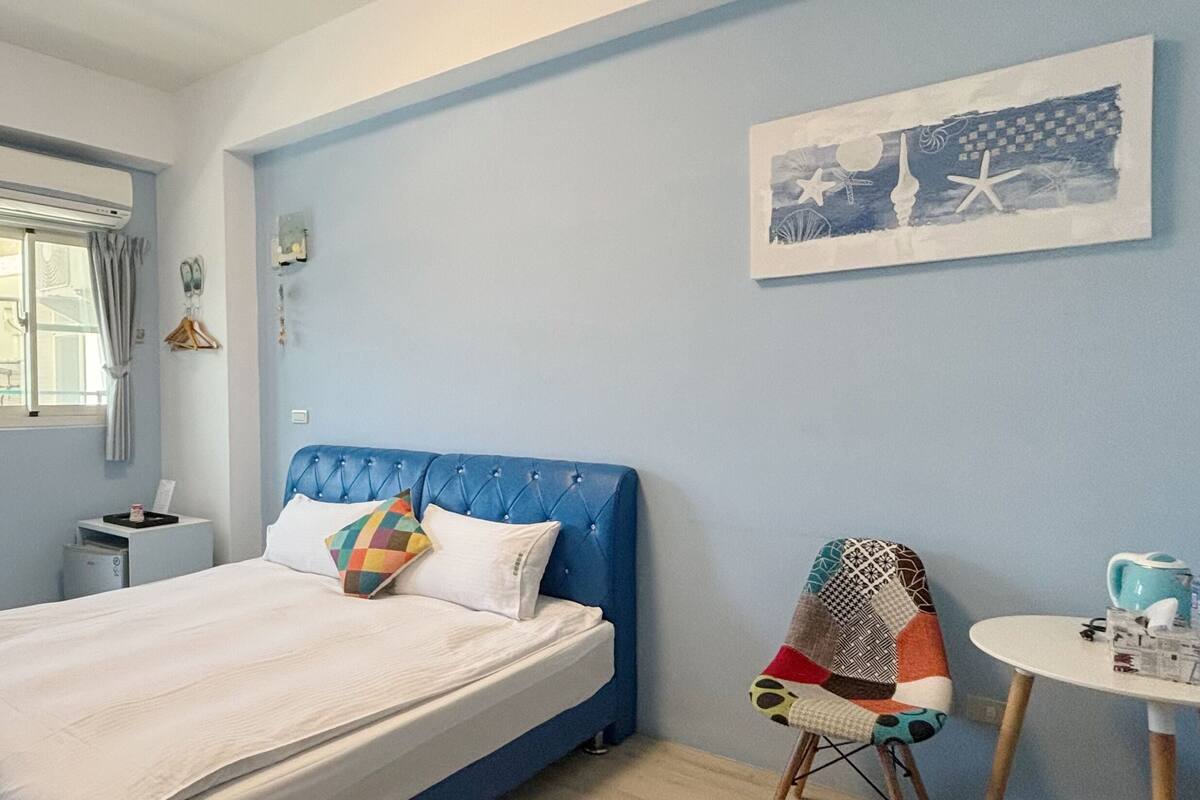 A bedroom with a bed, a chair, a small table, and a wall-mounted artwork.