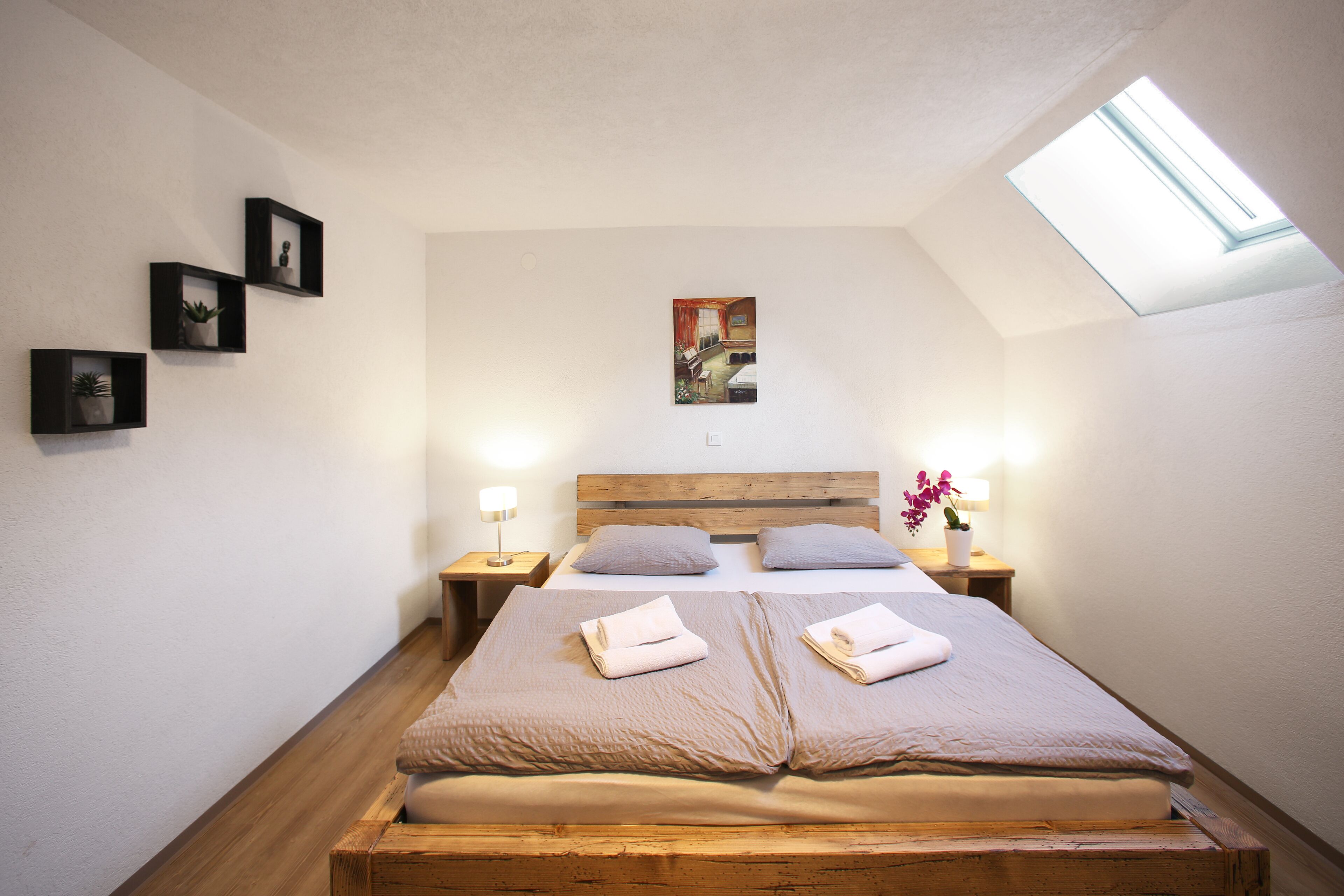 Une chambre avec une lucarne, un lit avec deux oreillers, une table de chevet avec une lampe et trois tableaux encadrés accrochés au mur.