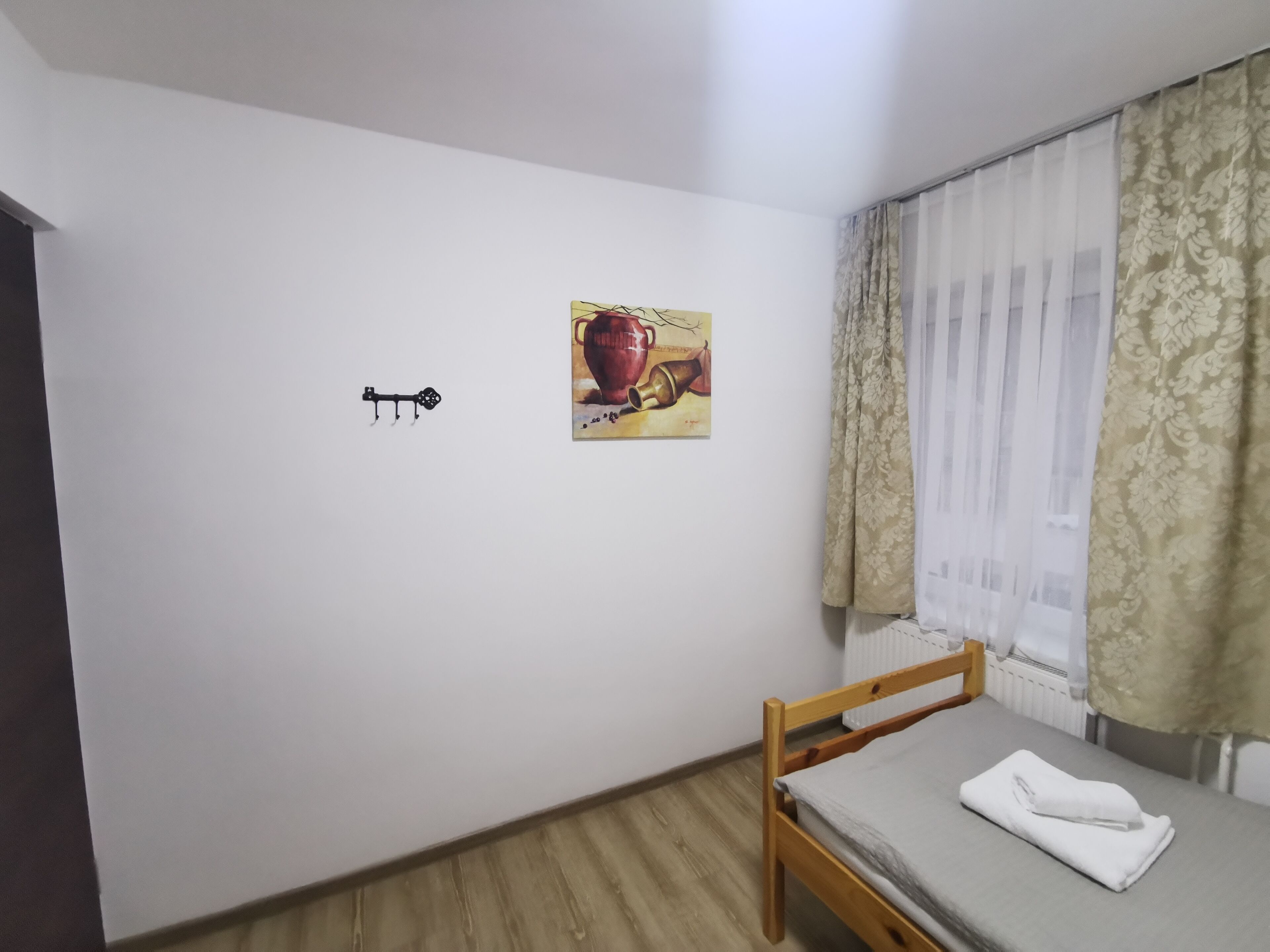 A small, tidy room with a wooden bed, a white wall with a single artwork, and a window with curtains.