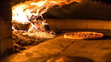 A pizza oven with flames, a pizza in the process of baking, and a pizza on a peel ready to be cooked.