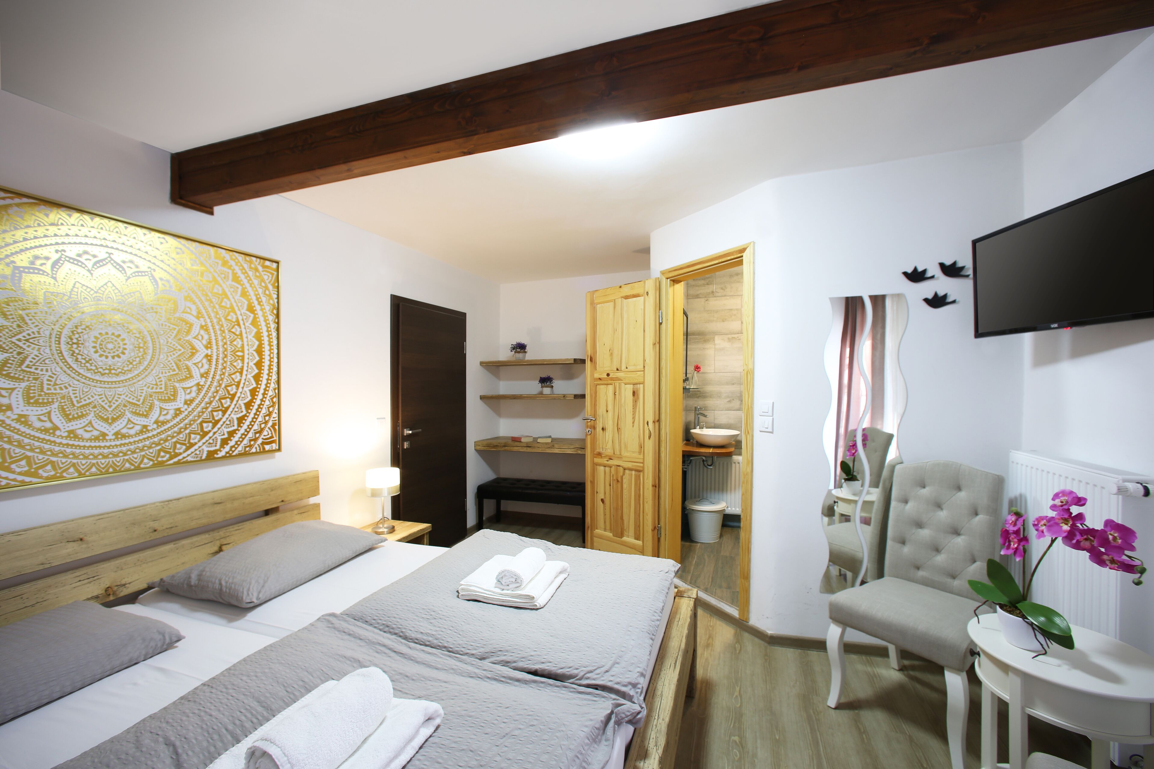 A hotel room with a large bed, a wooden headboard, a mirror, a television, and a small table with a flower vase.