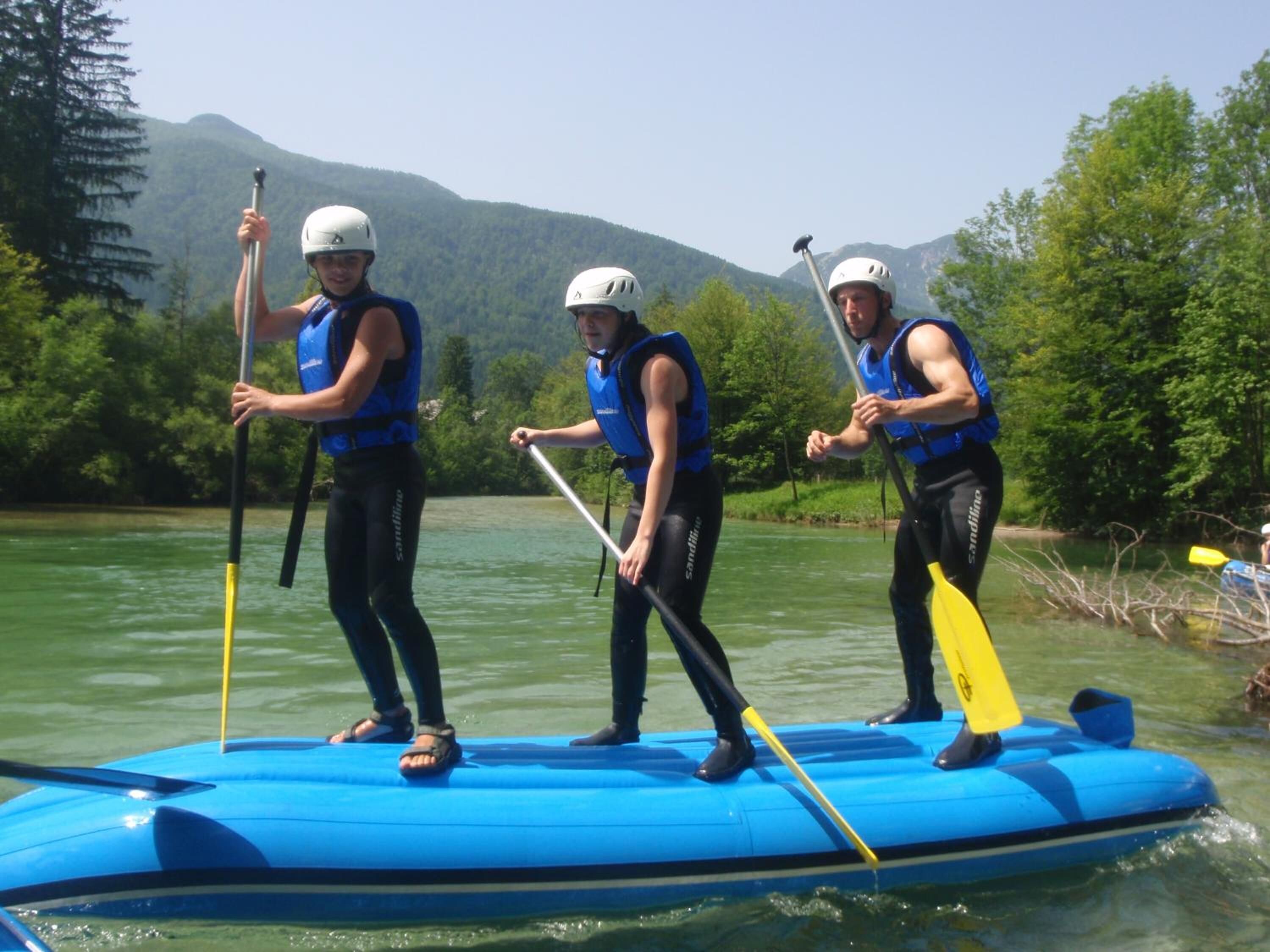 Three people on a blue raft paddleboarding on a river with mountains in the background.
