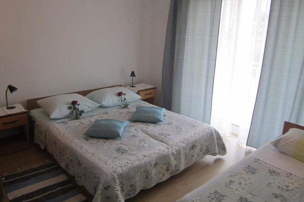 A bedroom with a large bed, a smaller bed, a nightstand, a lamp, and a window with curtains.