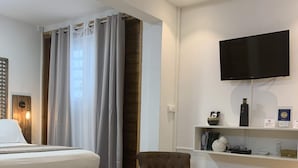 Une chambre d’hôtel avec un lit, un fauteuil marron, une table basse ronde, un téléviseur fixé au mur et une étagère contenant divers objets.