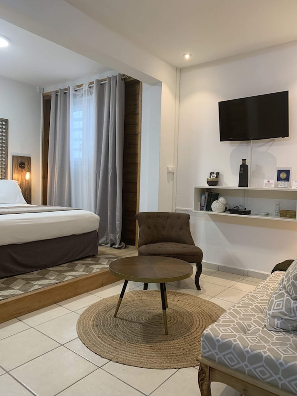 Une chambre d’hôtel avec un lit, un fauteuil marron, une table basse ronde, un téléviseur fixé au mur et une étagère contenant divers objets.